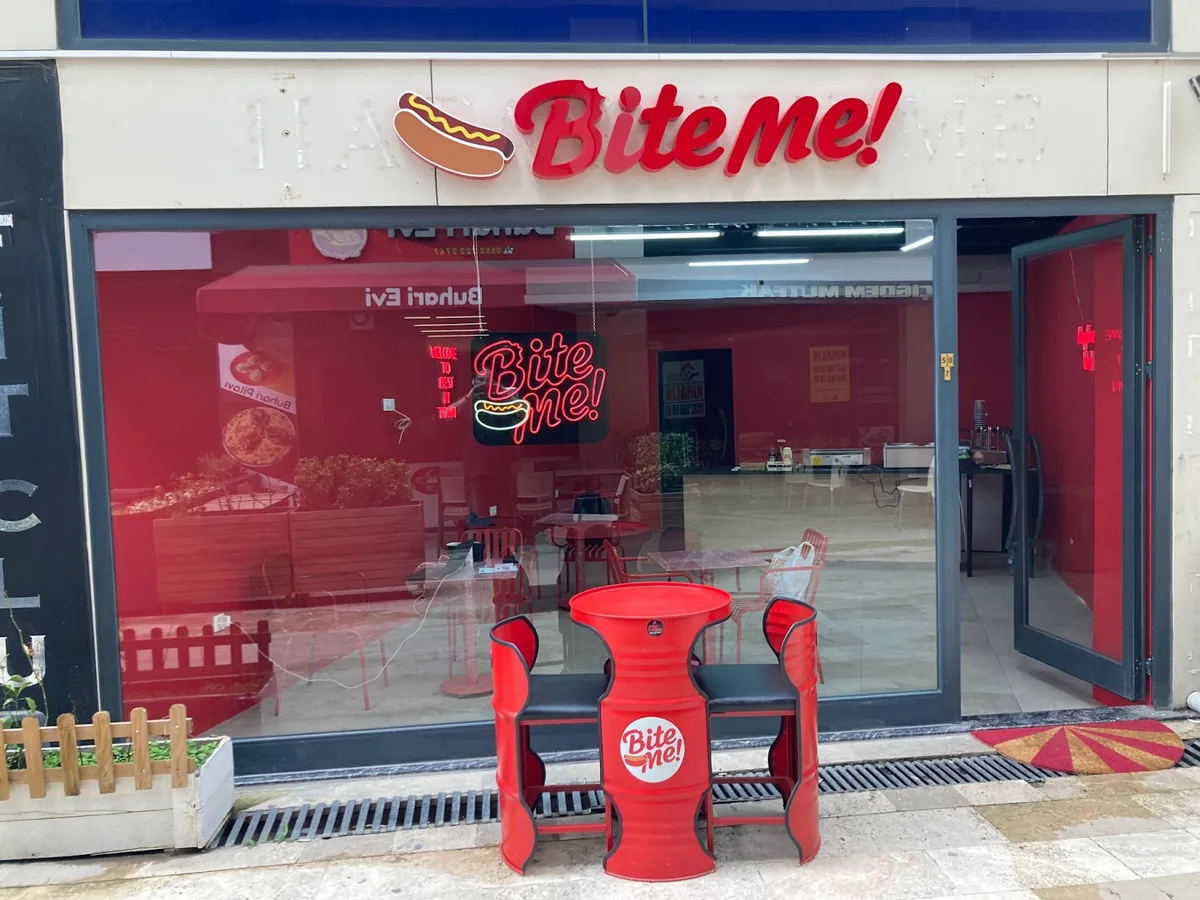 Bite Me! restoranının vitrini: kırmızı tabela, neon logo ve önünde logolu varil masa